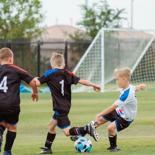 Three children playing soccer on a grass field with a goal in background